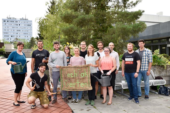 Gruppenfoto des TechFakGarten. 13 Personen, die mit Gießkannen und andere Werkzeuge zum Gärtnern bewaffnet vor der bepflanzten Betonrinne auf dem Roten Platz der TechFak stehen.