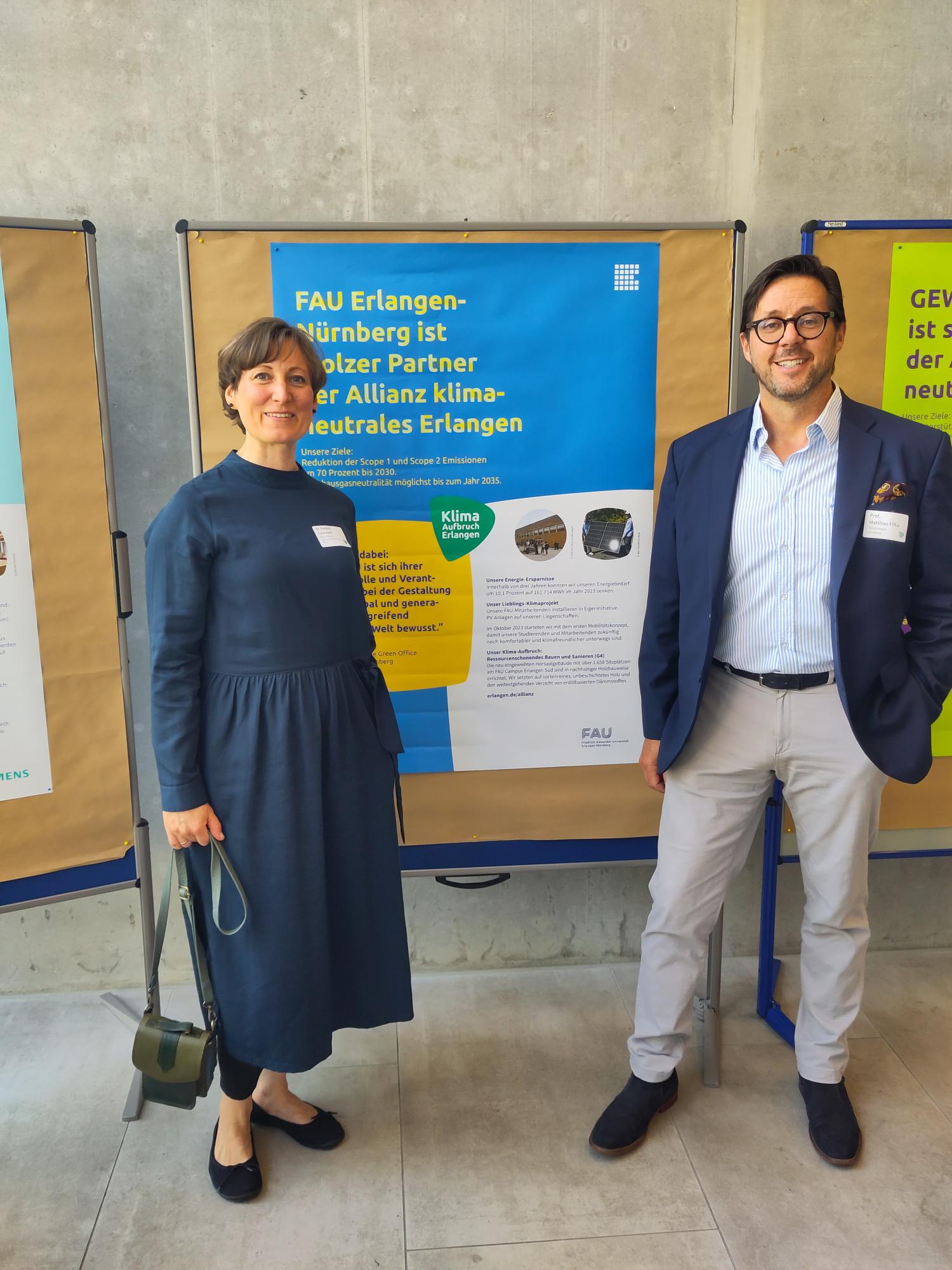 Foto zweier Personen vor einem Plakat mit dem Titel 