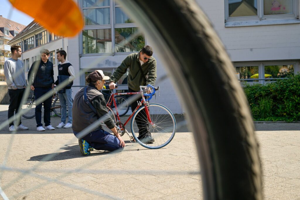 Zu sehen ist eine Gruppe an Menschen, die sich ein Fahrrad anschauen. Im Vordergrund des Fotos sind unscharf die Speichen eines Reifens zu sehen.
