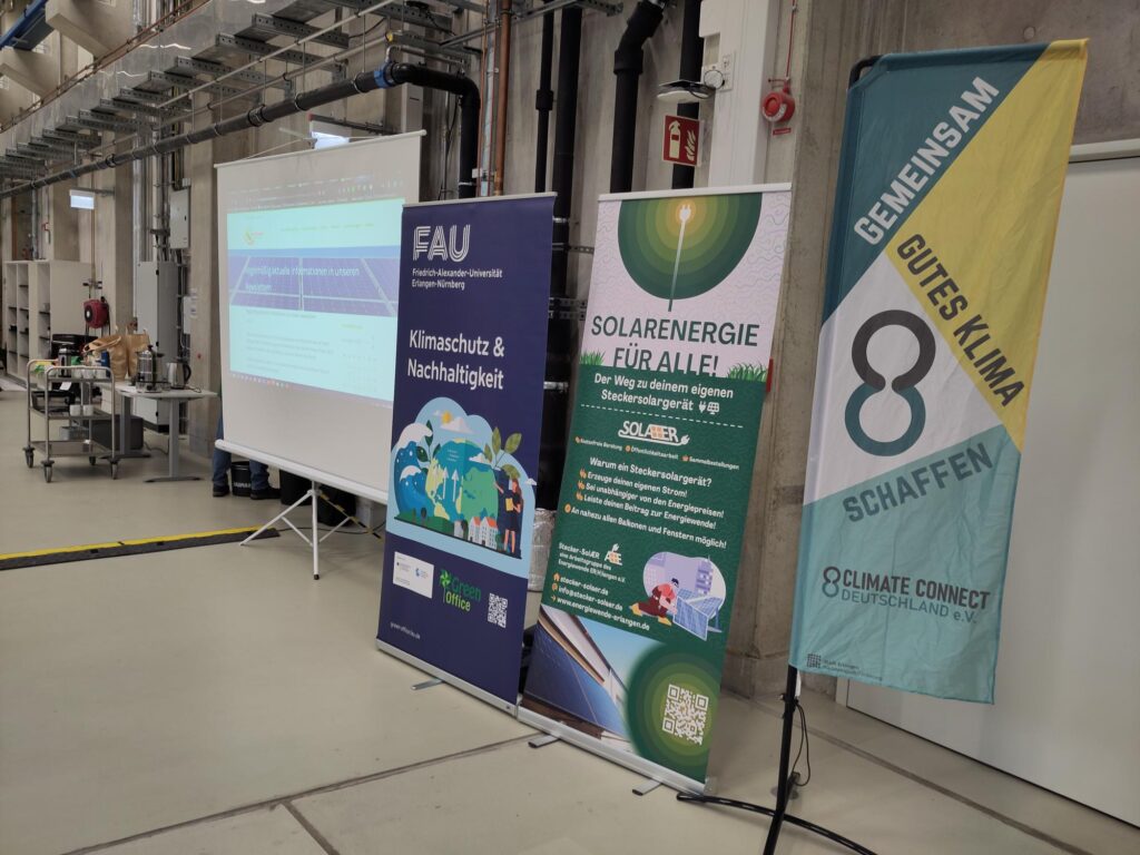 Auf dem Foto ist eine mobile Leinwand zu sehen, auf der eine Präsentation gezeigt wird. Außerdem ist ein Rollup des Green Office, der Initiative Stecker Sola(e)r und von ClimateConnect zu sehen.