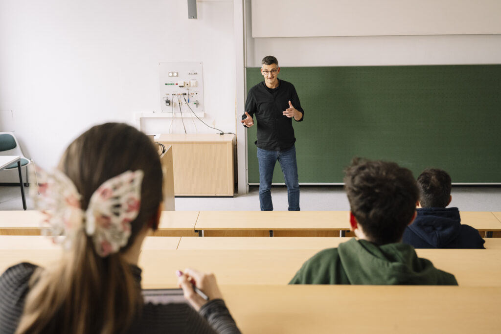 Auf dem Foto ist ein Dozierender zu sehen, der in einem Hörsaal vor der Tafel steht und gerade etwas erklärt. Außerdem sind die Hinterköpfe von drei Studierenden abgebildet, die gerade in den Stuhlreihen sitzen.