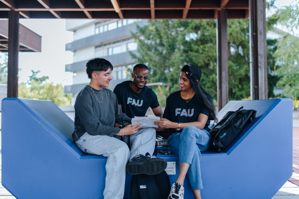 Auf dem Foto sind drei Studierende zu sehen, die auf einer Sitzgelegenheit im Freien sitzen und gemeinsam ein Blattpapier anschauen und darüber sprechen. Zwei Studierende tragen ein T-Shirt mit FAU-Logo.