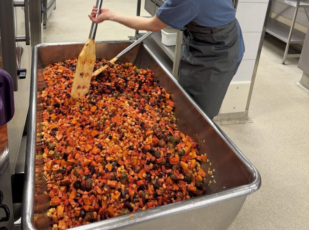 Auf dem Foto ist eine große Wanne, gefüllt mit kleingeschnittenem Gemüse zu sehen. Eine Person rührt das Gemüse gerade mit einem großen Spatel um.