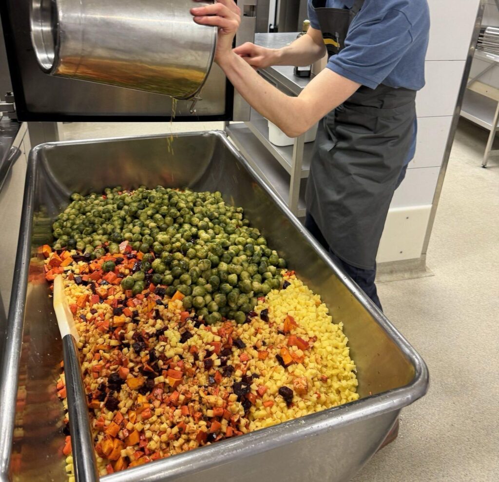 Auf dem Foto ist eine große Wanne, gefüllt mit kleingeschnittenem Gemüse zu sehen. Eine Person leert gerade noch weiteres Gemüse mit einem Eimer hinein.