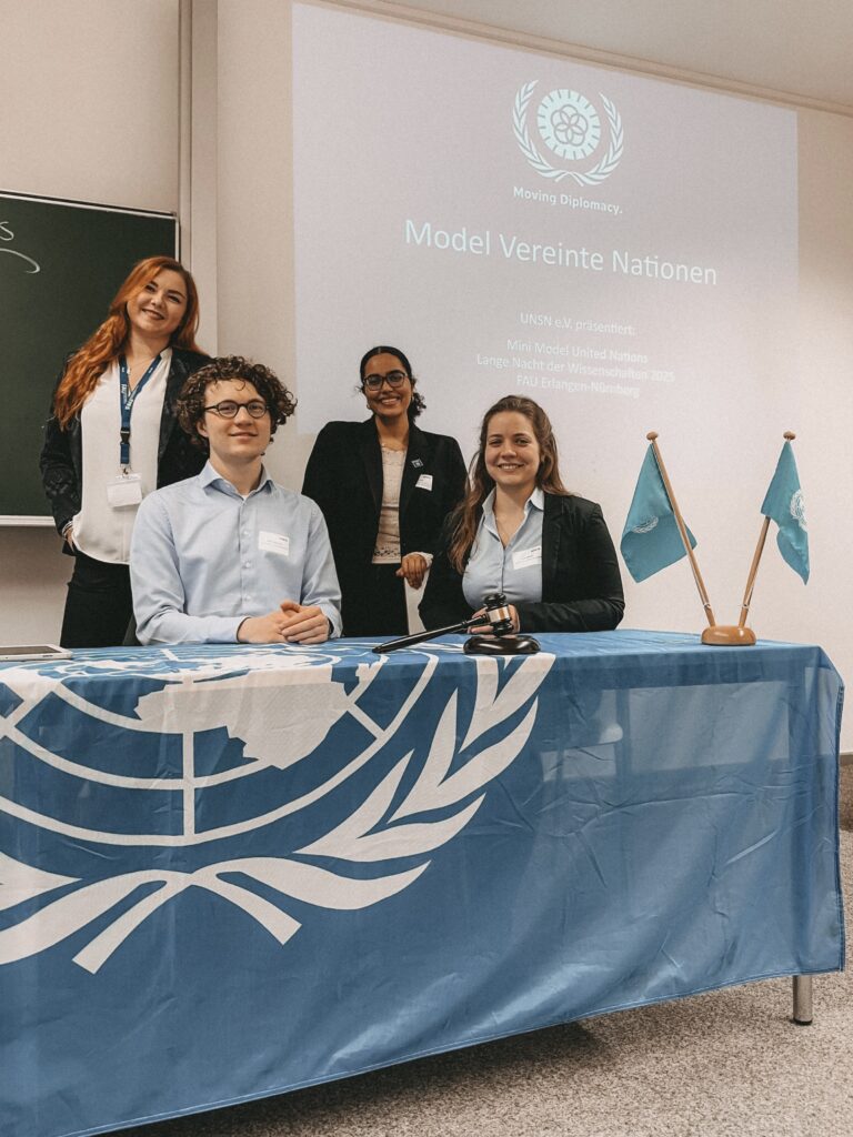 Auf dem Foto sind vier Personen hinter einemTisch stehend und sitzend zu sehen. Der Beamer strahlt eine Folie an die Wand, auf der "Model Vereinte Nationen" steht.