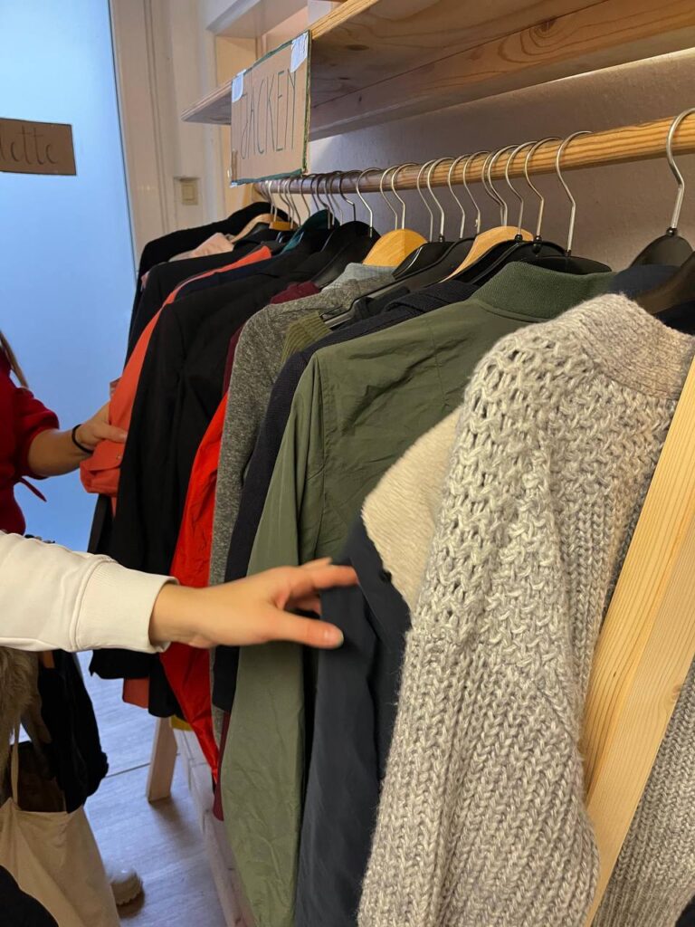 Auf dem Foto ist ein Kleiderständer mit Jacken zu sehen und die Arme von zwei Personen, die sich die Jacken anschauen.
