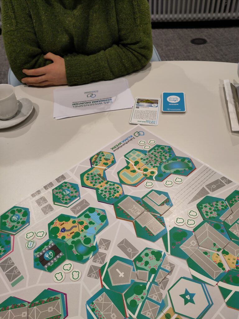 Im Vordergrund ist ein Spielbrett zui sehen mit vielen bunten Spielsteinen. Im Hintergrund sitzt eine Person in einem grünen Pullin mit einer weißen Tasse auf der linken Seite.