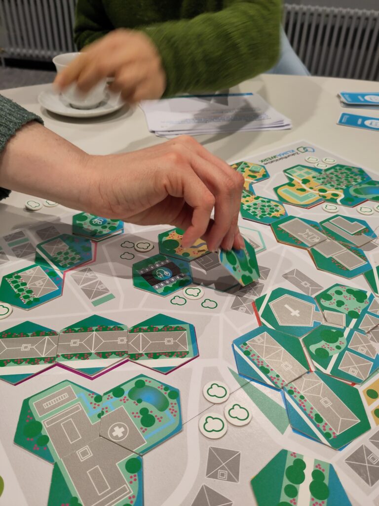 Eine Hand legt von der rechten Seite des Bildes einen Spielstein auf das Spielfeld. Im Hintergrund hebt eine Person in einem grünen Pulli einen Spielstein mit Baumsymbol auf.