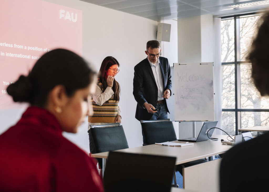 Auf dem Foto ist ein Dozierender abgebildet, der einer weiteren Person gerade etwas am Birldschirm zeigt. Im Vordergrund ist unscharf das Profil einer Person zu sehen. Im Hintergrund ist eine Präsentation an der Wand zu sehen.