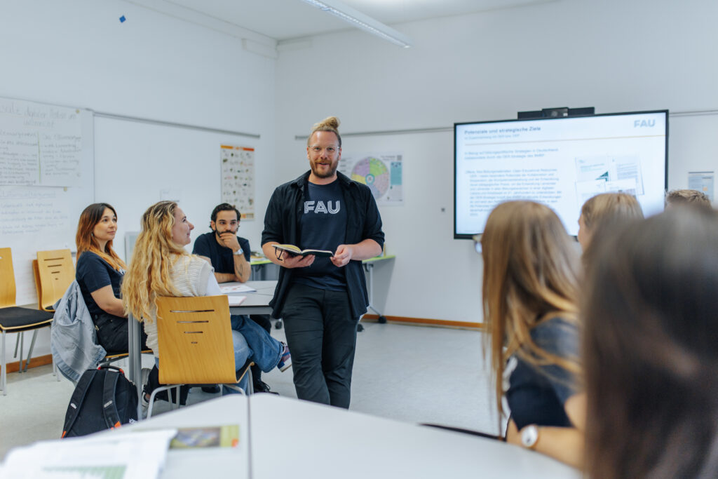 Zu sehen sind interessiert zuhörende Studierende und ein Dozent in der Mitte, der ein Buch in der Hand hält und lächelt. Im Hintergrund ist ein Bildschirm mit einer Präsentationsfolie unscharf zu erkennen.