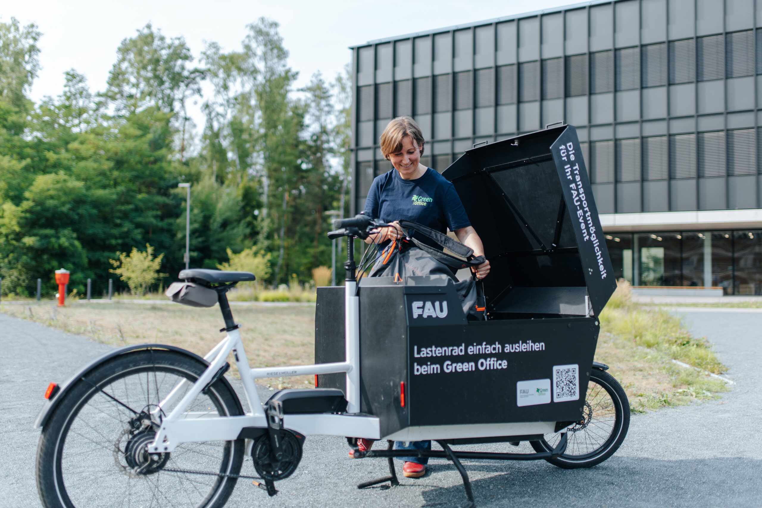 Auf dem Foto ist das E-Lastenrad des Green Office abgebildet. Die Transportbox ist beklebt mit dem FAU-Logo sowie dem Text "Lastenrad einfach ausleihen beim Green Office". Eine Frau stellt gerade eine Tasche in die Transportbox.