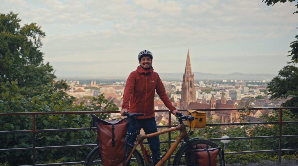 Auf dem Foto ist Ingwar Perowanowitsch, ein junger Mann und Aktivist, zu sehen. Er lächelt glücklich, trägt einen Fahrradhelm und eine rote Regenjacke und steht hinter seinem Fahrrad steht. An dem Fahrrad sind Satteltaschen vorne und hinten befestigt. Er steht vor einem Geländer und im Hintergrund ist ein Kirchturm zu sehen.