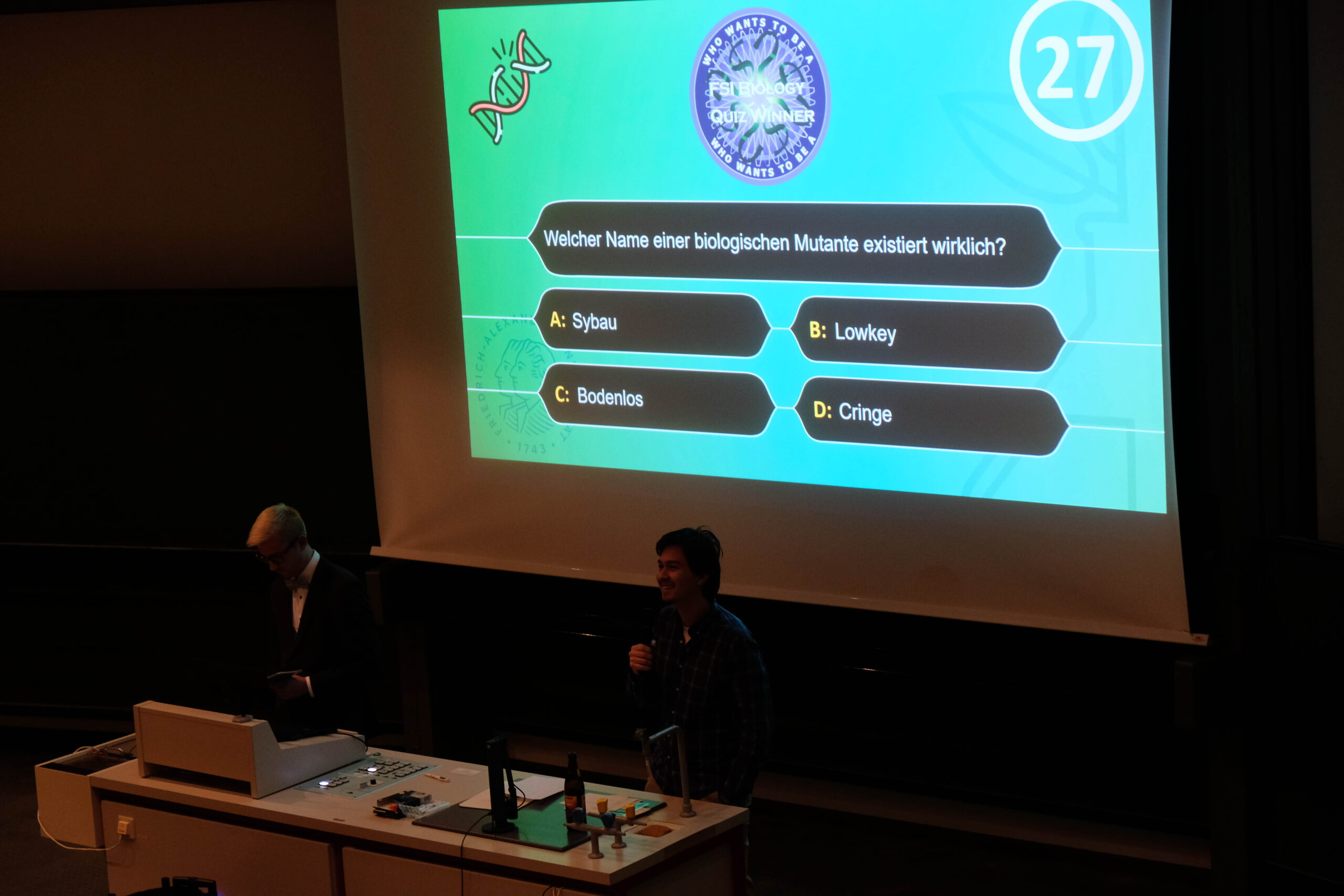 Zu sehen ist eine Präsentation mit einer Quiz-Frage und 2 Personen, die die Fragen moderieren mit einem Mikrophon.