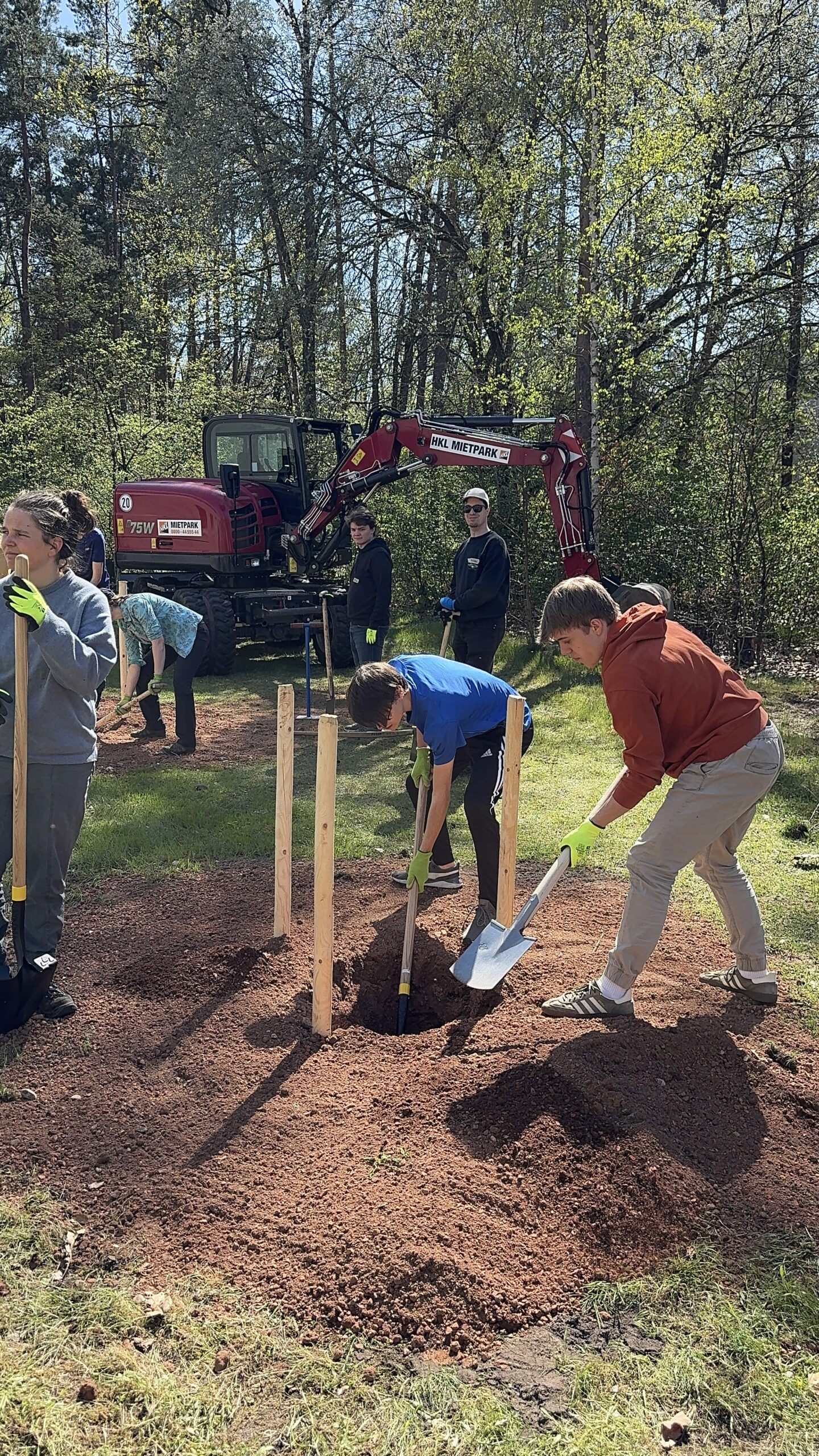 Im Vordergrund schaufeln zwei Studies ein Loch. Im Hintergrund steht ein Bagger.